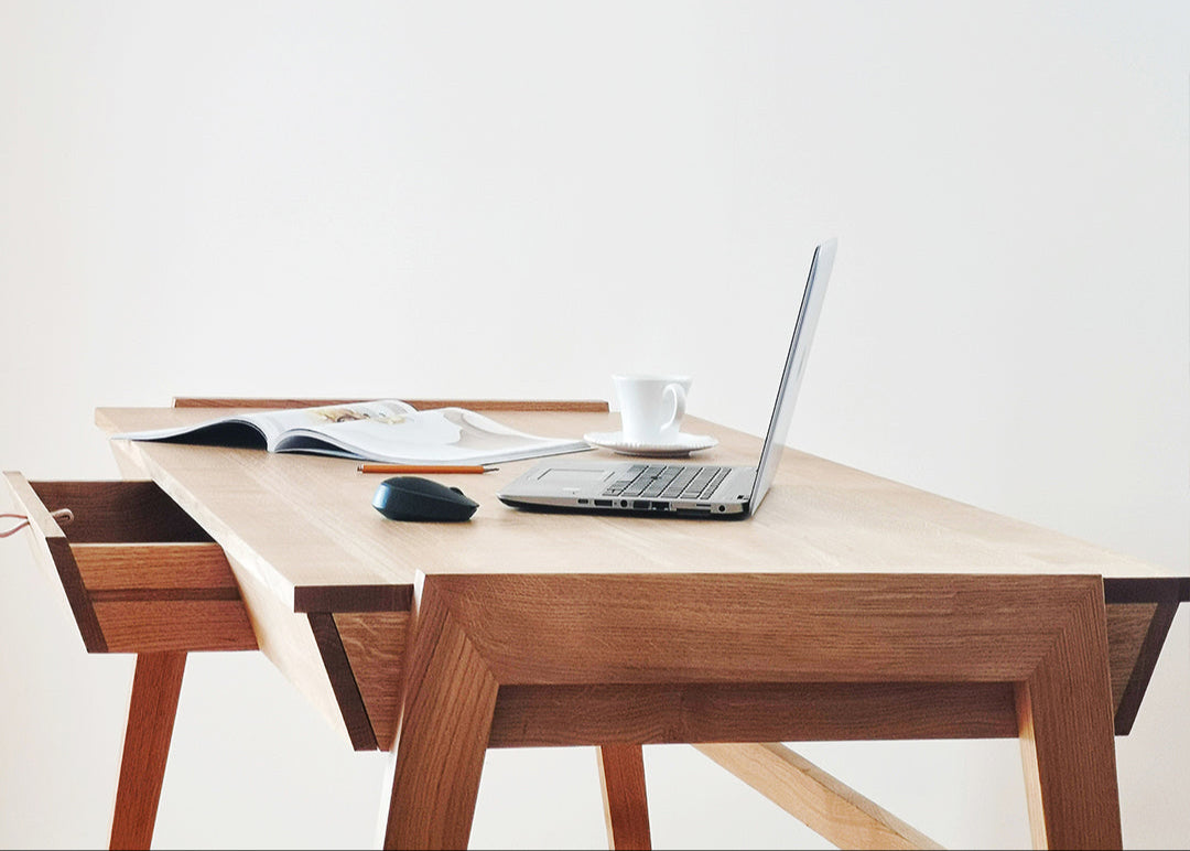 The Solid Oak Desk with Drawer - Model_No.01 by KUNSST FURNITURE provides a minimalist display for a laptop, an open magazine, a mouse, and a white coffee cup against a plain white background.