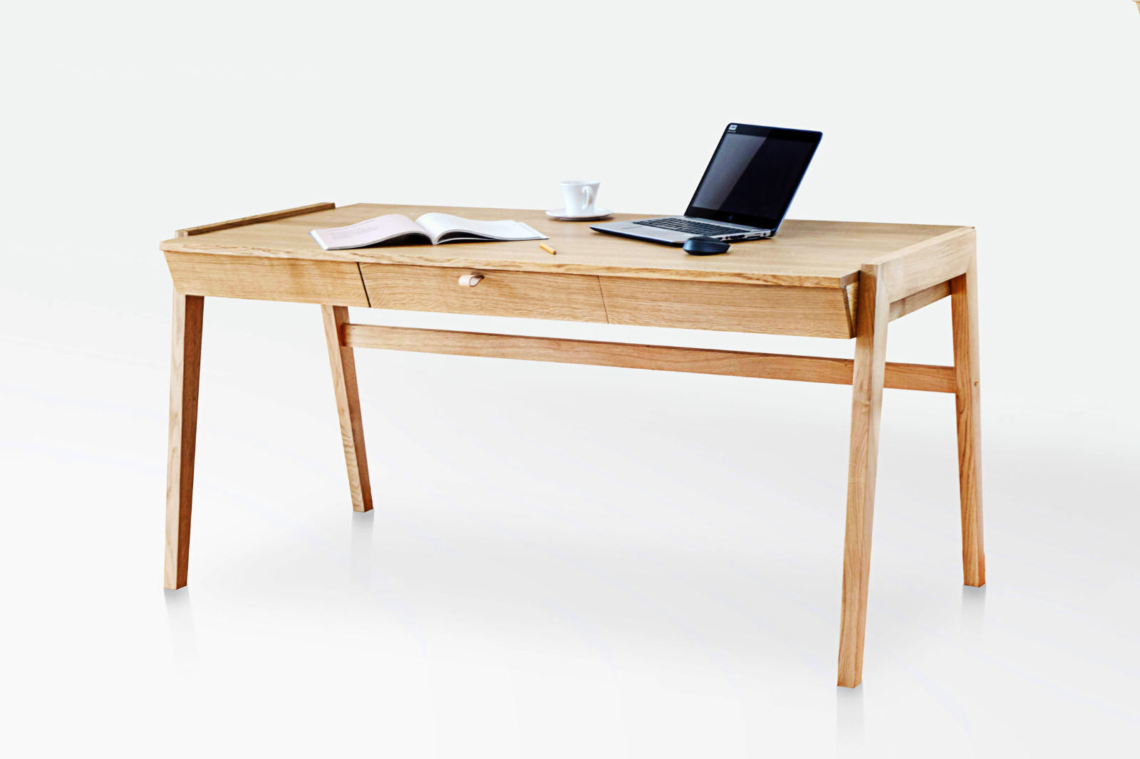 Minimalist oak desk with a central drawer and angled legs, with a book, cup and laptop on top