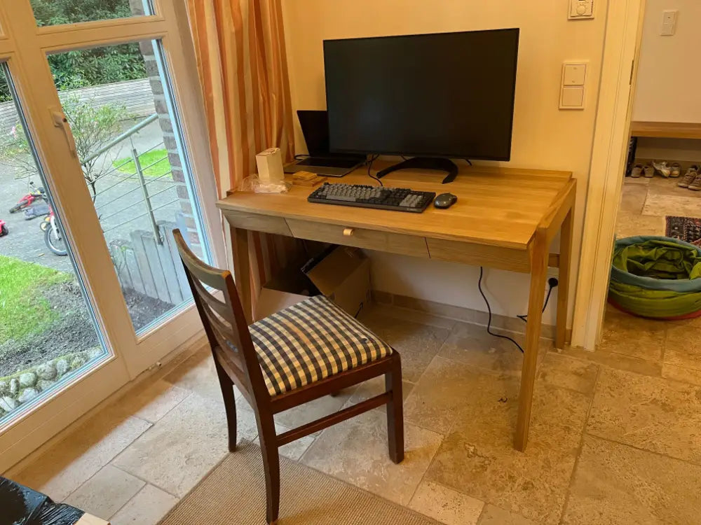 View of solid wood desk with drawer with computer setup in a room with large windows.