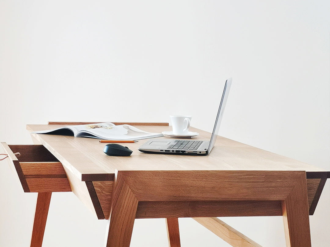 The Solid Oak Desk with Drawer - Model_No.01 by KUNSST FURNITURE provides a minimalist display for a laptop, an open magazine, a mouse, and a white coffee cup against a plain white background.