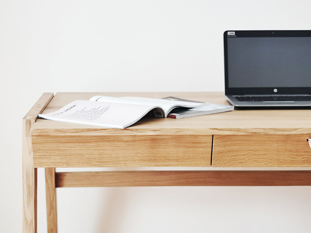 A minimalist workspace is showcased with the KUNSST FURNITURE Solid Oak Desk with Drawer - Model_No.01, featuring a handcrafted drawer slightly open to display an open magazine and a laptop, all against a plain white wall, highlighting the clean and modern aesthetics inspired by traditional woodworking techniques.