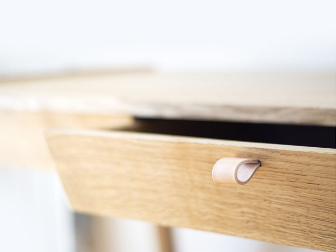 A close-up of the Solid Oak Desk with Drawer - Model_No.01 by KUNSST FURNITURE highlights its small leather pull handle on a partially open drawer. This desk, crafted with traditional woodworking techniques, features a light, natural finish against a softly blurred background.