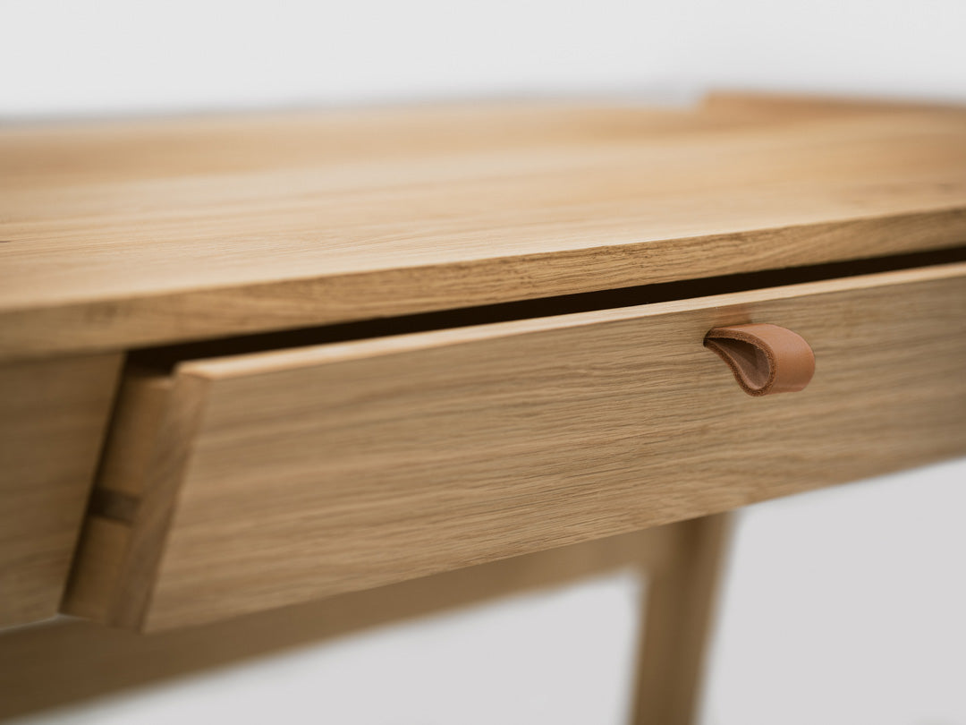 Close-up of the Solid Oak Desk with Drawer - Model_No.01 by KUNSST FURNITURE, showcasing a partially open drawer with a simple leather pull handle. This solid oak desk displays a natural wood finish, while the background is out of focus, emphasizing its minimalist design and traditional woodworking craftsmanship.