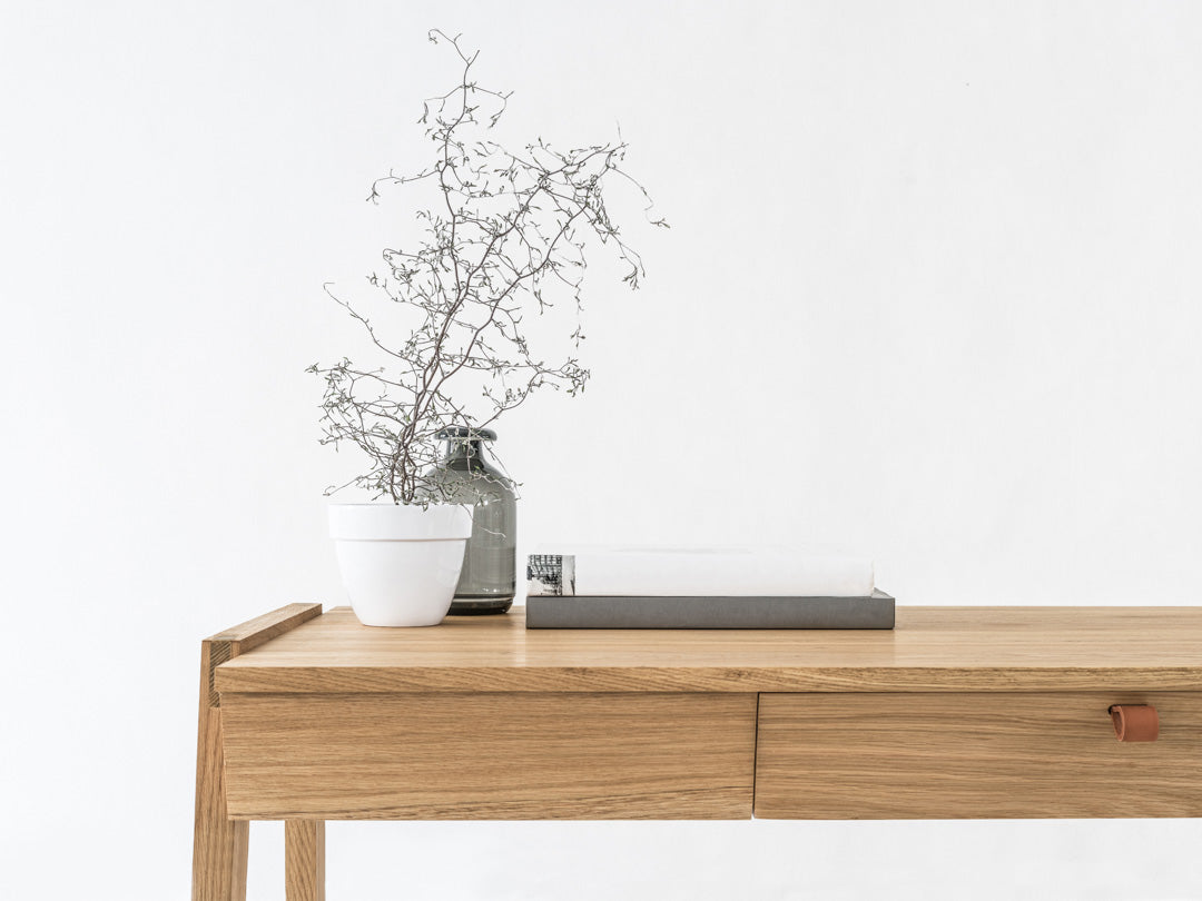 The KUNSST FURNITURE Solid Oak Desk with Drawer - Model_No.01 is a minimalist piece crafted using traditional woodworking techniques. It includes two handcrafted drawers and is complemented by a potted plant in a white pot, a stack of books, and a small glass vase holding delicate branches, all set against the simplicity of a plain white wall.