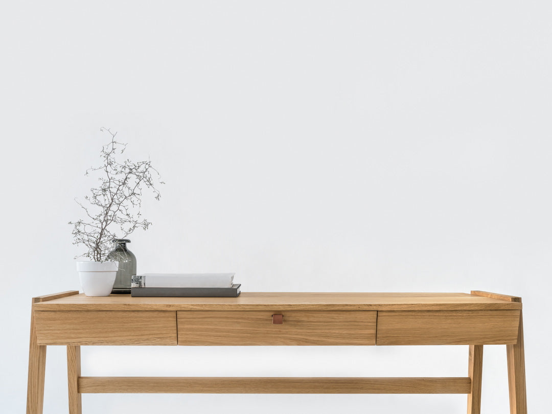 A Solid Oak Desk with Drawer - Model_No.01 by KUNSST FURNITURE sits against a plain white wall. On the left side, there is a vase containing a dried branch and a small white planter. A gray tray holds some papers on the surface. This desk, crafted using traditional woodworking techniques, features one handcrafted central drawer.