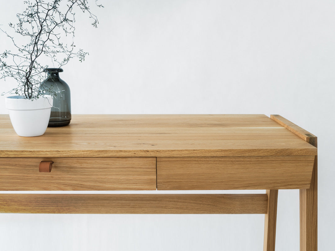 The Solid Oak Desk with Drawer - Model_No.01 by KUNSST FURNITURE showcases a minimalist design featuring two handcrafted drawers, one adorned with a leather handle. Atop the desk sits a dark glass vase containing a dried branch and an elegant white pot. The plain white backdrop accentuates the simplicity of the design while highlighting the charm of traditional woodworking techniques.