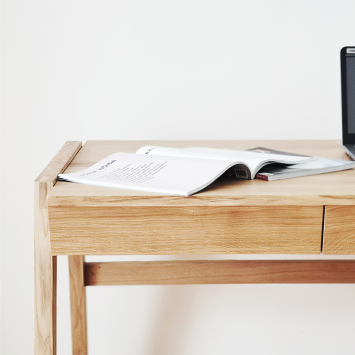 A KUNSST FURNITURE Solid Oak Desk with Drawer - Model_No.01 is gracefully positioned against a plain white wall, adorned with an open magazine and a partially visible laptop. It highlights traditional woodworking techniques, featuring clean lines and a light natural finish, complemented by a handcrafted drawer for added elegance.