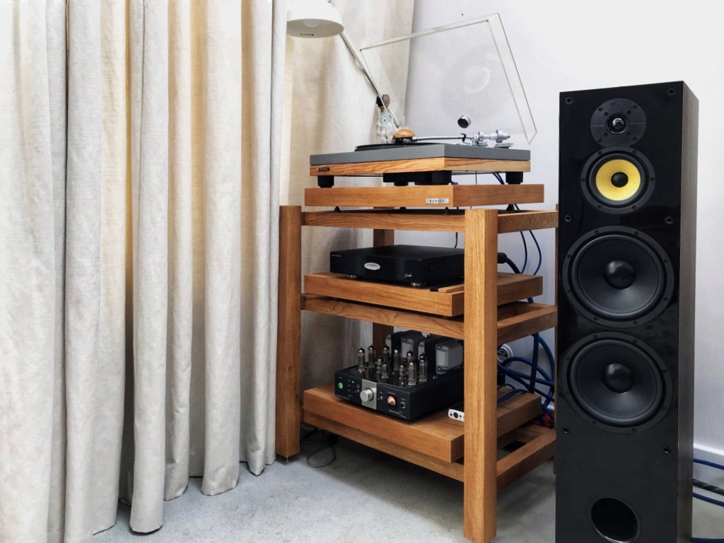 Dreistöckiges Plattenspieler Rack in einer Wohnzimmerecke vor einem weichen Vorhang, oben ein Victrola Montauk Plattenspieler, in der Mitte eine Fezz Audio Gratia Phono-Vorstufe und unten ein Tsakiridis Hermes Röhrenverstärker